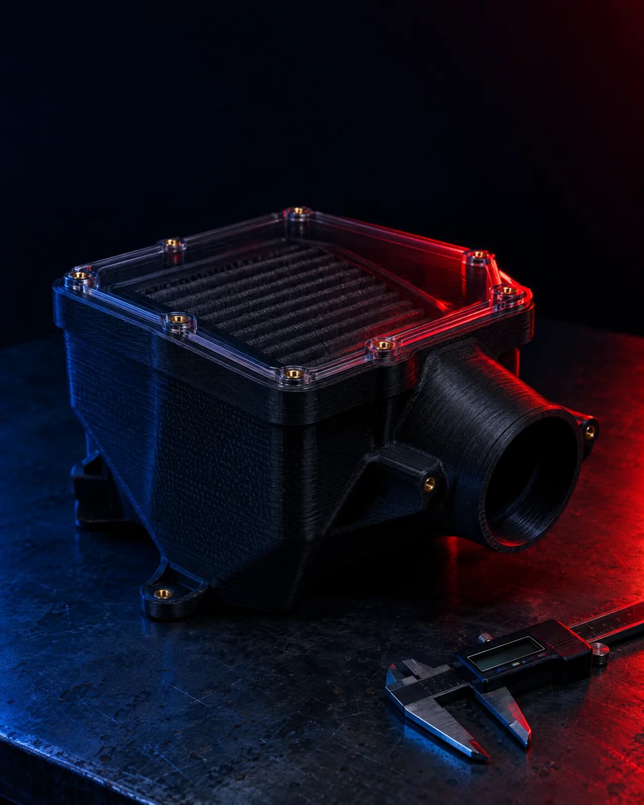 Black carbon-fibre 3D-printed airbox with a clear lid and brass inserts, on a steel bench beside vernier calipers