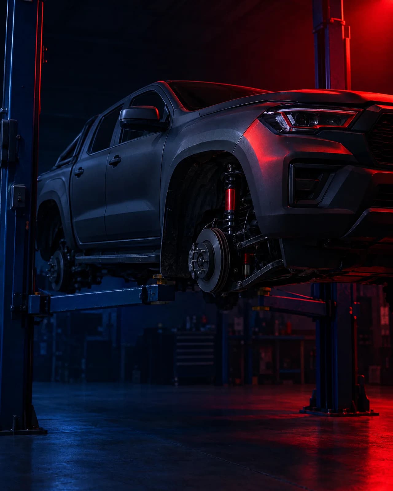 A dark grey dual-cab ute on a workshop hoist with new dampers fitted, under a red key light